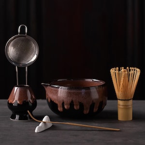 Puede incluir: Un juego de té matcha que incluye un cuenco de cerámica esmaltada en negro y marrón, un soporte para batidor a juego con un colador de acero inoxidable, un batidor de bambú y una cuchara de bambú sobre un soporte de cerámica blanca.