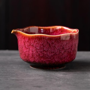 May include: A ceramic tea pitcher with a vibrant magenta and deep red crystalline glaze. The vessel features a wide, wavy rim and a small, integrated pouring spout. The exterior surface displays a textured, mottled pattern with dark undertones.