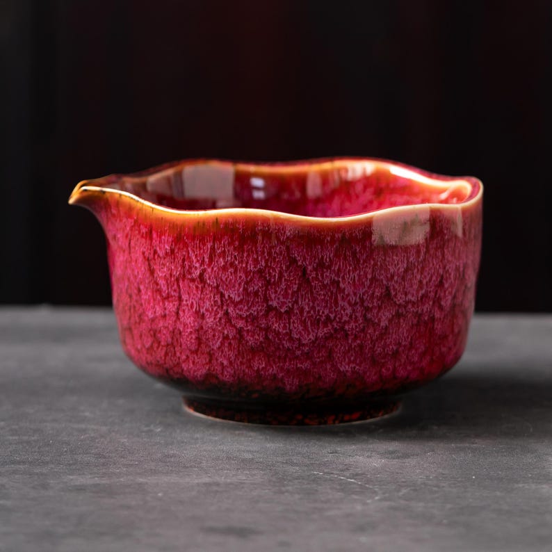 May include: A ceramic tea pitcher with a vibrant magenta and deep red crystalline glaze. The vessel features a wide, wavy rim and a small, integrated pouring spout. The exterior surface displays a textured, mottled pattern with dark undertones.