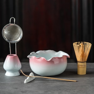 Op de afbeelding: Een matcha theeset met een keramische kom en gardehouder met een roze en witte gradiëntafwerking, een bamboe garde, een metalen zeef en een dunne bamboe lepel die rust op een kleine witte keramische standaard in de vorm van een wolk.