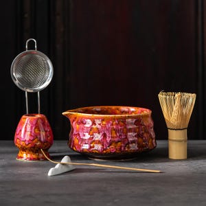 Puede incluir: Un juego de té matcha con un cuenco de cerámica, un batidor, un colador y un pequeño soporte de cerámica. El cuenco y el soporte del colador tienen un acabado texturizado y esmaltado en tonos naranja, rosa y blanco. El batidor es de bambú.