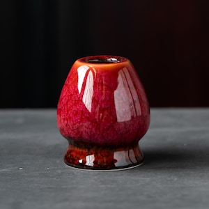 May include: A ceramic matcha whisk holder featuring a glossy, vibrant red glaze with a mottled, textured finish. The base of the holder transitions into a deep, dark brown tone, providing a distinct contrast to the bright red upper section.