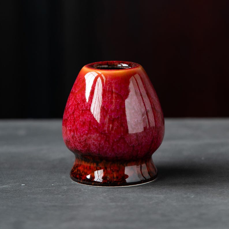 May include: A ceramic matcha whisk holder featuring a glossy, vibrant red glaze with a mottled, textured finish. The base of the holder transitions into a deep, dark brown tone, providing a distinct contrast to the bright red upper section.