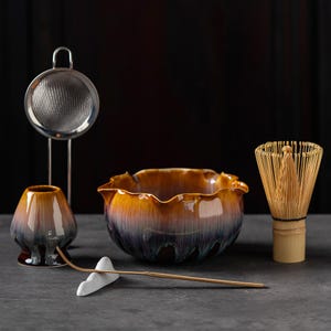 May include: A matcha tea set featuring a ceramic bowl with a scalloped rim and a gradient glaze of amber, purple, and dark brown. The set includes a bamboo whisk, a ceramic whisk holder, a metal mesh strainer, and a bamboo scoop on a white rest.