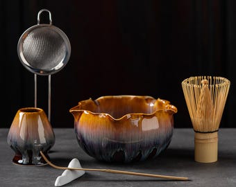 May include: A matcha tea set featuring a ceramic bowl with a scalloped rim and a gradient glaze of amber, purple, and dark brown. The set includes a bamboo whisk, a ceramic whisk holder, a metal mesh strainer, and a bamboo scoop on a white rest.