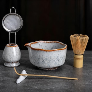 May include: A matcha tea set featuring a white and blue speckled ceramic bowl with a brown rim, a matching ceramic whisk holder with a metal mesh strainer, a bamboo whisk, and a long bamboo tea scoop resting on a white ceramic rest.