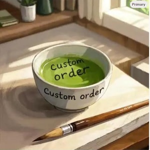 Puede incluir: Un cuenco de cerámica blanca lleno de líquido verde vibrante, con el texto "custom order" escrito en negro en el borde exterior y dentro del propio líquido. Un pincel de madera marrón se encuentra en diagonal en primer plano.