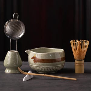 May include: A matcha tea set including a light green ceramic bowl with a brown dachshund illustration, a bamboo whisk, a bamboo scoop on a white rest, a green ceramic whisk holder, and a stainless steel mesh strainer on a metal stand.