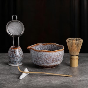 May include: A matcha tea set featuring a ceramic bowl and a matching whisk holder with a speckled blue and brown glaze, a bamboo whisk, a bamboo scoop, and a metal mesh strainer. The bowl has a pouring spout and a wavy rim design.