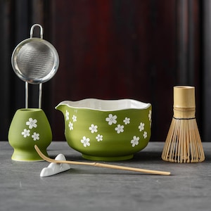 May include: A matcha tea set featuring a green ceramic bowl with white floral designs, a bamboo whisk, a stainless steel strainer, and a bamboo scoop. The set includes a white ceramic whisk holder. The bowl measures approximately 15 cm in diameter.