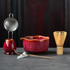 Peut inclure: Un ensemble à thé matcha comprenant un bol en céramique rouge profond avec un bec verseur, un porte-fouet rouge assorti avec une passoire en métal, un fouet en bambou et une fine cuillère en bambou posée sur un support blanc en forme de montagne.