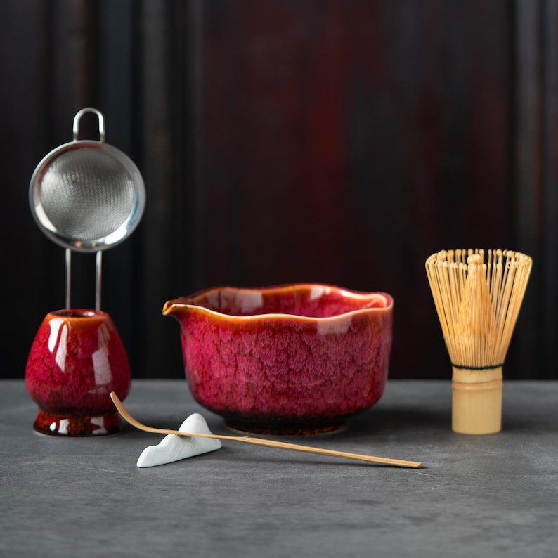 May include: A matcha tea set featuring a deep red ceramic bowl with a pour spout, a matching red whisk holder with a metal sieve, a bamboo whisk, and a thin bamboo scoop resting on a white ceramic mountain-shaped holder for tea preparation.