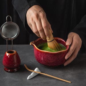 May include: A deep red ceramic matcha bowl with a pour spout, a bamboo whisk, a matching red ceramic whisk holder, a metal mesh strainer, and a long bamboo tea scoop resting on a white ceramic holder, used for preparing green tea.