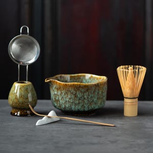 May include: A matcha tea set featuring a ceramic bowl with a teal and brown mottled glaze, a bamboo whisk, a bamboo scoop resting on a white mountain-shaped holder, and a metal mesh strainer sitting atop a matching ceramic whisk stand.