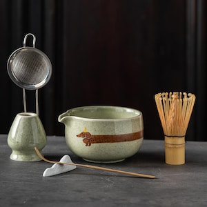 May include: A matcha tea set featuring a light green speckled ceramic bowl with a brown dachshund illustration, a bamboo whisk, a bamboo scoop on a white mountain-shaped rest, a ceramic whisk holder, and a stainless steel mesh strainer.