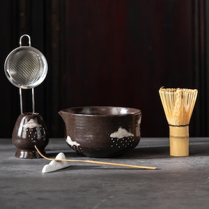 May include: A matcha tea set. It includes a dark brown ceramic bowl with cloud and raindrop designs, a bamboo whisk, a ceramic tea scoop rest, a tea strainer, and a holder. The set is for preparing matcha tea.