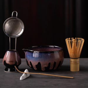 May include: A ceramic matcha bowl with a purple, blue, and black drip glaze, a matching whisk holder, a stainless steel mesh strainer, a bamboo whisk, and a long bamboo scoop resting on a small white ceramic stand against a dark background.
