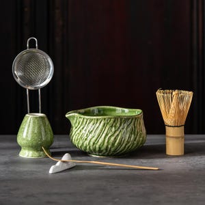 May include: A matcha tea set featuring a green ceramic bowl with a textured design, a bamboo whisk, a stainless steel strainer, and a ceramic whisk holder. The set is arranged on a dark surface, ideal for tea preparation.