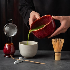 May include: A matcha tea set including a red glazed ceramic pouring bowl, a white ceramic bowl, a bamboo whisk, a bamboo scoop on a white rest, and a metal mesh strainer on a red glazed ceramic stand. Green matcha tea is being poured.