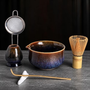 May include: A matcha tea set featuring a ceramic bowl with a blue and brown gradient glaze, a bamboo whisk, a bamboo scoop resting on a white ceramic holder, and a metal whisk stand with an attached fine mesh strainer.