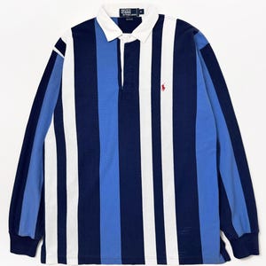May include: A long-sleeved rugby shirt with vertical stripes in navy, white, and light blue. The shirt has a white collar, a three-button placket, and a small red logo on the chest. The shirt is made of a soft, comfortable material.