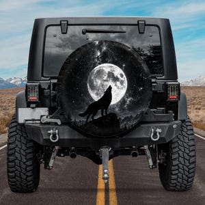 May include: A black spare tire cover mounted on the rear of a black off-road vehicle. The cover features a circular graphic of a wolf howling at a large, bright full moon set against a dark, starry night sky with silhouettes of mountain peaks.