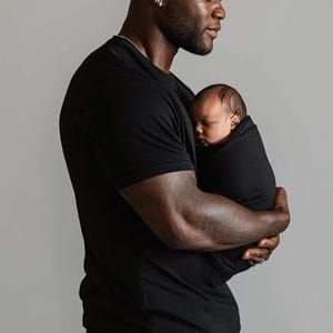 Pu&ograve; includere: Una maglietta nera tiene in braccio un bambino fasciato. Il bambino &egrave; avvolto in un tessuto nero, con solo il viso visibile. La persona che tiene il bambino indossa una maglietta nera e un orecchino d'argento.