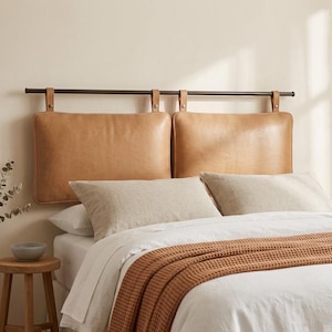 May include: A bed with a headboard featuring two tan leather cushions suspended from a dark metal bar. The bed is made with white linens, a brown waffle-knit blanket, and decorative pillows. A small wooden stool and a decorative plant are in the foreground.