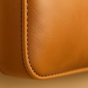 May include: Close-up of a section of a tan leather bag. The leather has a smooth texture with visible grain. The edges are neatly stitched with a contrasting thread, showcasing the craftsmanship and design of the bag.