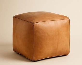 May include: A square, brown leather ottoman. The ottoman is a cube shape with stitched seams and a smooth, slightly textured surface. The color is a warm, medium brown.