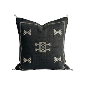 May include: A square, dark gray decorative pillow with a geometric pattern. The pillow features a central rectangular design and four X-shaped patterns, all in a lighter gray. The edges are trimmed with a lighter gray stitching and tassels.