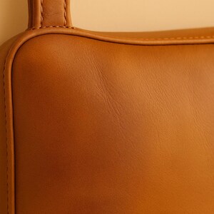 May include: Close-up of a section of a brown leather bag. The leather has a smooth, slightly textured surface with visible grain. The bag features a rounded corner and a stitched handle.