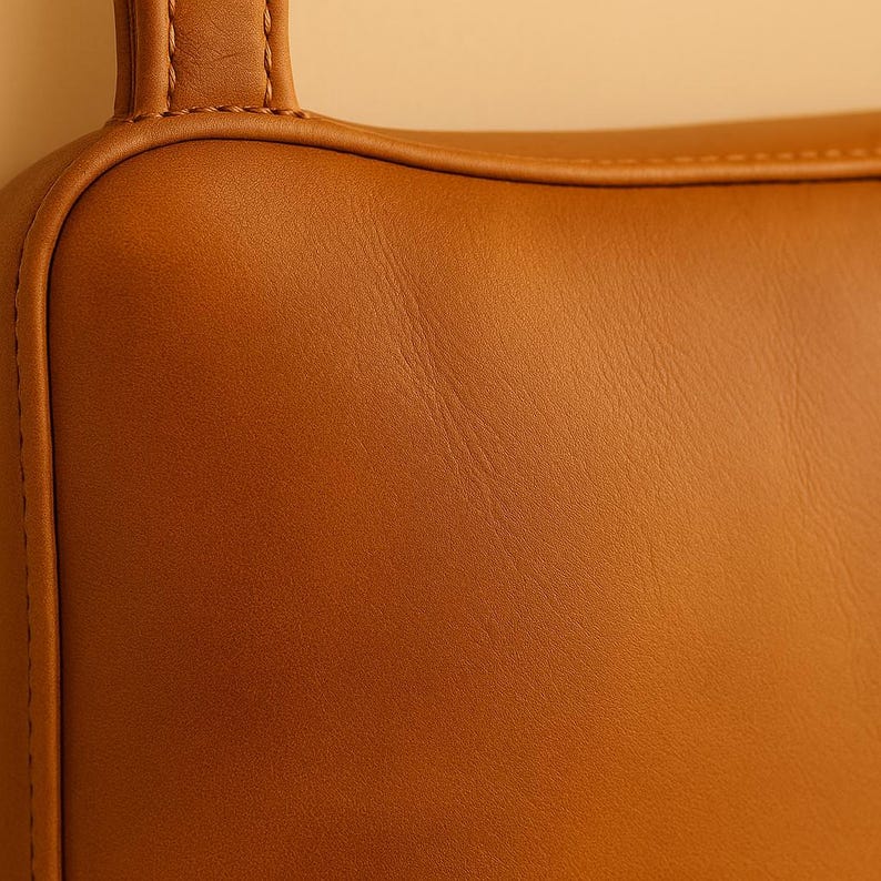 May include: Close-up of a section of a brown leather bag. The leather has a smooth, slightly textured surface with visible grain. The bag features a rounded corner and a stitched handle.