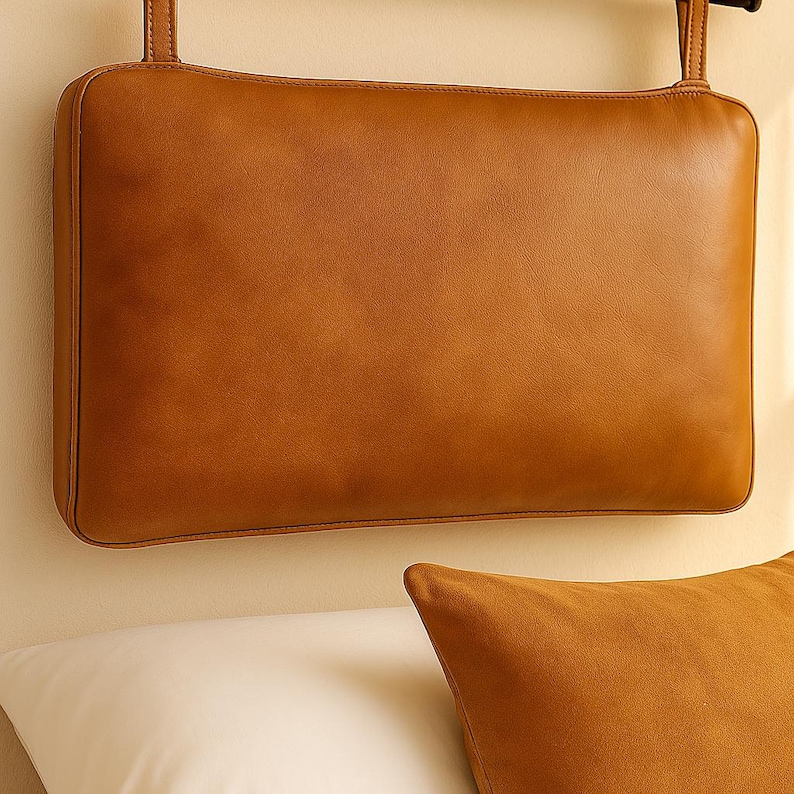 May include: A rectangular, tan leather headboard with two hanging straps. The headboard is a solid color and has a smooth texture. A matching tan pillow is in the foreground, resting on a white pillow.