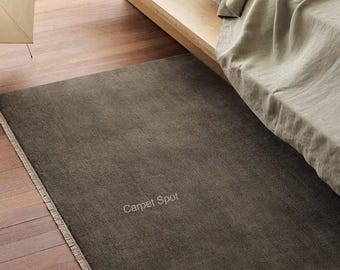 Pode incluir: Um tapete retangular marrom escuro com uma borda com franjas, colocado em um piso de madeira. O texto "Carpet Spot" é visível na superfície do tapete. O tapete está posicionado perto de uma cama com um cobertor de cor clara.