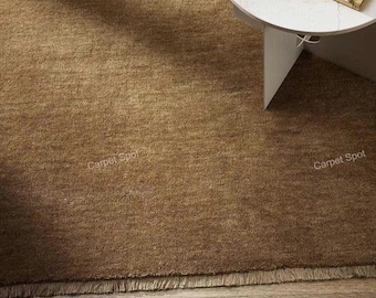 May include: A close-up of a brown rug with a textured surface and a fringed edge. The rug is made of a soft material and is placed on a wooden floor. A white round table is in the background.