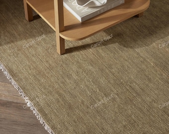 May include: A textured, light brown area rug with a woven pattern and white fringe along the edge. A light-colored wooden side table sits on the rug, holding a white, wavy-edged decorative bowl placed on top of a book titled Abode.