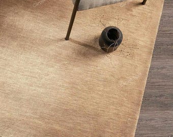 Puede incluir: Una alfombra rectangular beige con un borde con flecos, combinada con una silla moderna y un pequeño jarrón negro. La alfombra tiene un aspecto texturizado y está colocada sobre un suelo de madera. La silla tiene un asiento de color claro y patas de metal oscuro.