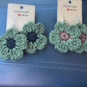 May include: Two pairs of handmade crochet flower earrings. One pair has green petals with a dark blue centre, the other has green petals with a mauve centre. Each pair is attached to a card that reads "Handmade with love."