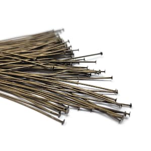 May include: A pile of antique brass-colored head pins on a white background. The pins have a slender, straight shaft and a flat, round head. The pins are used for jewelry making and other craft projects.