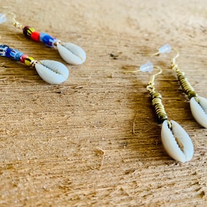 May include: A pair of gold earrings with cowrie shells and colorful beads. The earrings are on a wooden surface.