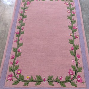 May include: A rectangular area rug featuring a solid pink centre bordered by a lavender frame. A continuous vine pattern with green leaves and small pink flowers with yellow centres runs along the inner perimeter of the rug, creating a floral border.