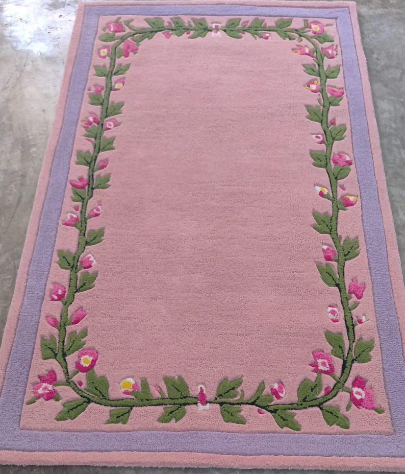 May include: A rectangular area rug featuring a solid pink centre bordered by a lavender frame. A continuous vine pattern with green leaves and small pink flowers with yellow centres runs along the inner perimeter of the rug, creating a floral border.