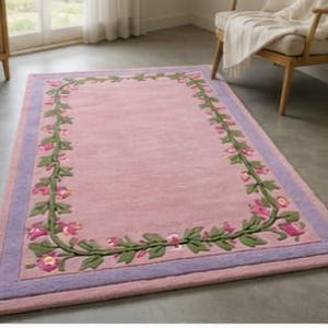 May include: A rectangular area rug featuring a solid pink centre field surrounded by a decorative border of green vines and pink flowers. The rug is framed by a wide lavender border and an outer pink edge, creating a layered, colourful design.