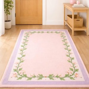 May include: A rectangular area rug featuring a solid light pink centre framed by a decorative border of green vines with small pink flowers. The rug is finished with a wide, solid lavender purple border along the outer edge of the design.