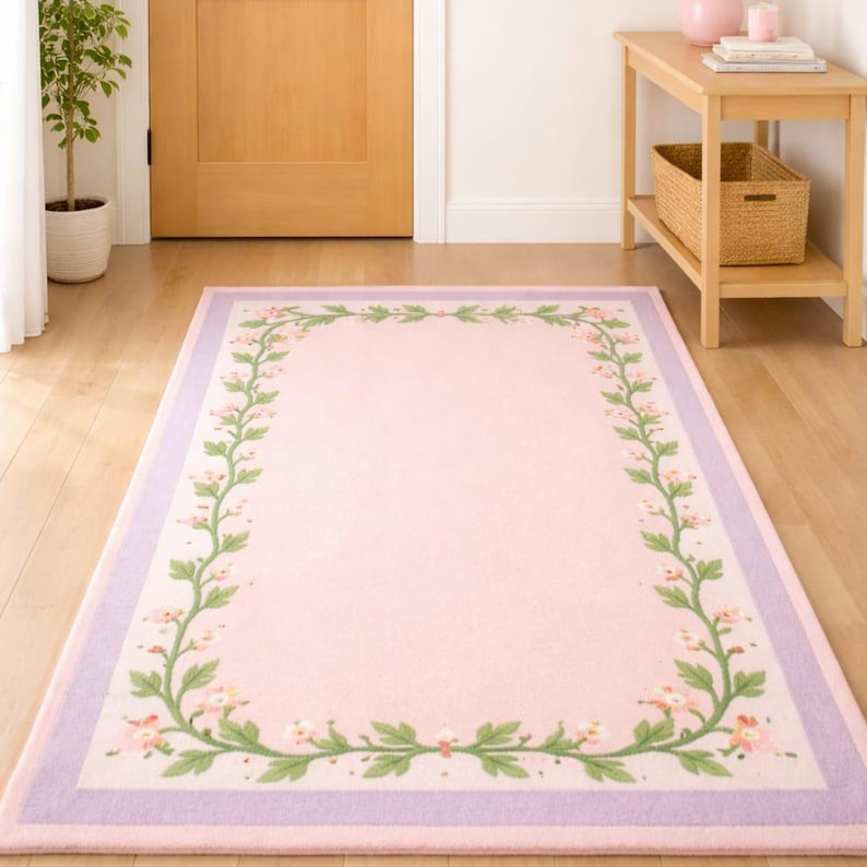 May include: A rectangular area rug featuring a solid light pink centre framed by a decorative border of green vines with small pink flowers. The rug is finished with a wide, solid lavender purple border along the outer edge of the design.