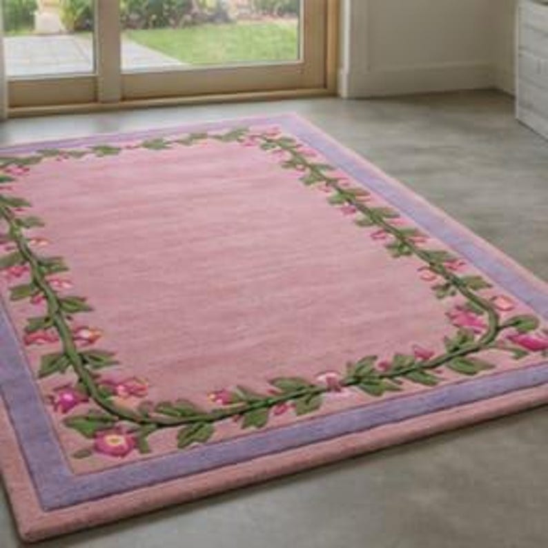 May include: A rectangular area rug featuring a solid pink centre field surrounded by a decorative border of green vines and pink flowers. The rug is framed by a light purple outer border, creating a distinct contrast against the pink interior.
