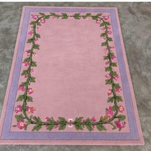 May include: A rectangular area rug featuring a solid light pink centre field framed by a decorative border of green vines with pink flowers. The rug is finished with a wide lavender border that surrounds the floral vine pattern on all four sides.
