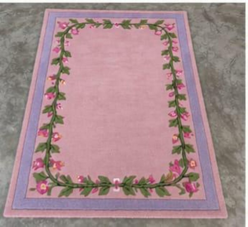 May include: A rectangular area rug featuring a solid light pink centre field framed by a decorative border of green vines with pink flowers. The rug is finished with a wide lavender border that surrounds the floral vine pattern on all four sides.