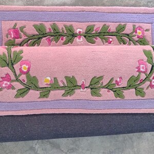 May include: A rectangular tufted rug featuring a soft pink centre with a green vine and pink flower motif, framed by a lavender border. The rug is folded over to reveal a dark grey textured backing on the underside of the fabric material.
